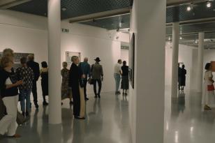 Układ poziomy. Przestrzeń galeryjna. Jasne podłogi i ściany. Kilkanaście osób oglądających wystawę.