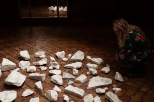 Fotografia barwna w układzie poziomym. białe odłamki kamienia, różnej wielkości rozrzucone na brązowym parkiecie. Rozrzucone fragmenty tworzą krąg oświetlony jasnym światłem.