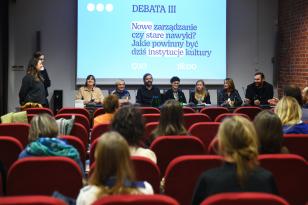 Fotografia pozioma. Duża sala z miękkimi krzesłami ustawionymi w rzędach jak w kinie, na ścianie w głębi na dużym ekranie wyświetlana prezentacja. Pod ekranem za stołem siedzi 7 osób, obok stoją 2 osoby. W fotelach siedzi kilkanaście osób.